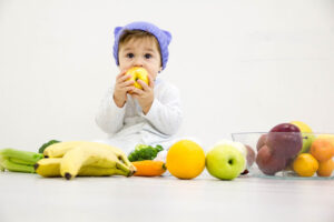 Toddler surrounded by fruit and vegetables, attempting to eat an apple.