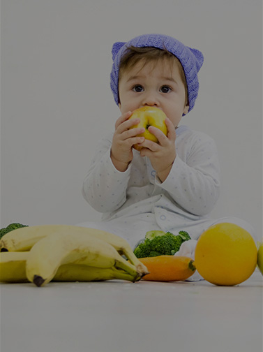 Toddler surrounded by fruit and vegetables, attempting to eat an apple.