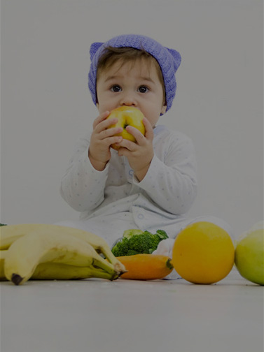 Toddler surrounded by fruit and vegetables, attempting to eat an apple.