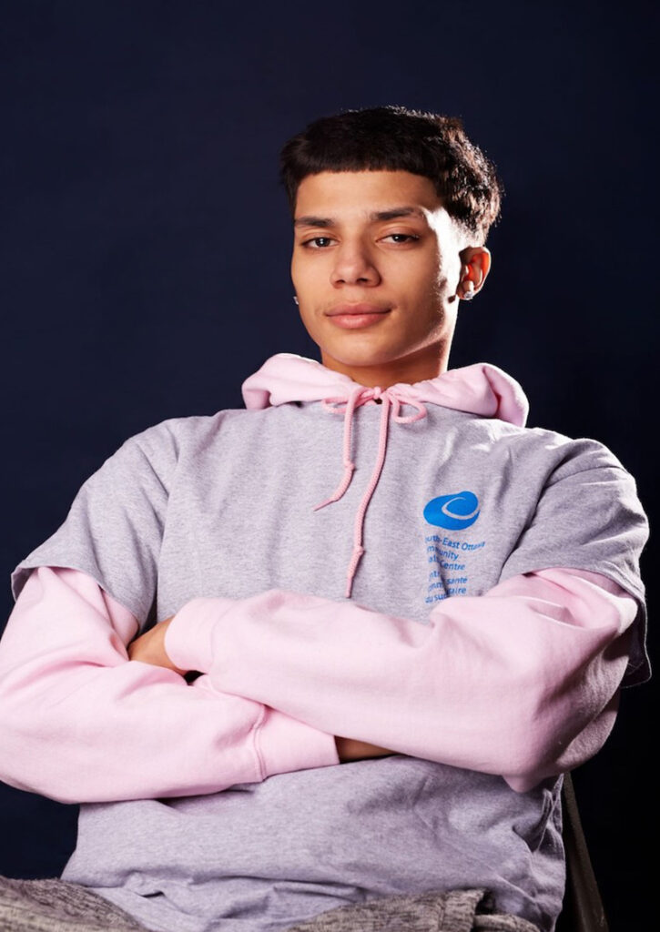 Boy with pink hoodie leaning back with arms folded.