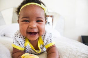 Smiling infant with yellow hair band laying on adult bed.