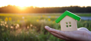 Hand holding tiny wooden house in foreground with sunrise over tree line in background.